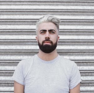 Close-up portrait of a man with a beard and gray hair against a striped metal background.